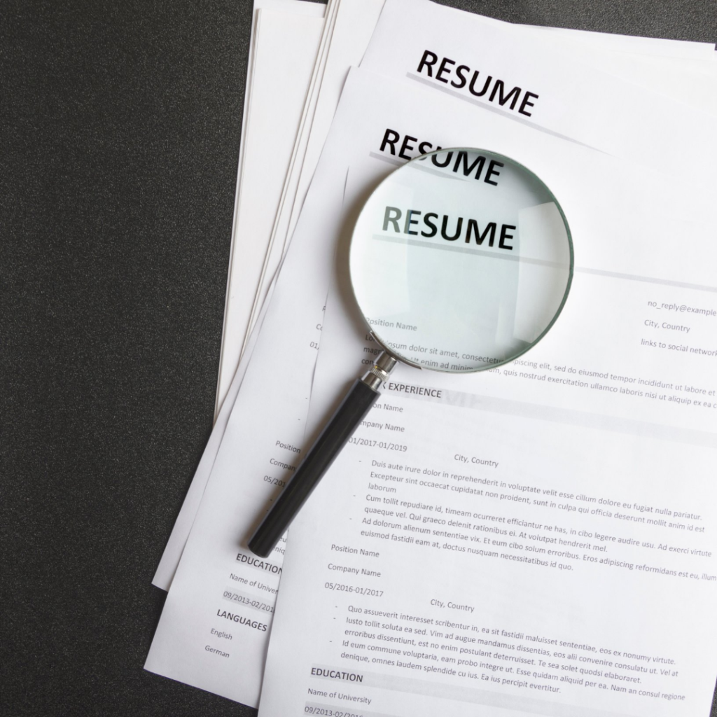 Several generic resumes stacked on top of each other with a magnifying glass magnifying the word 'resume'.